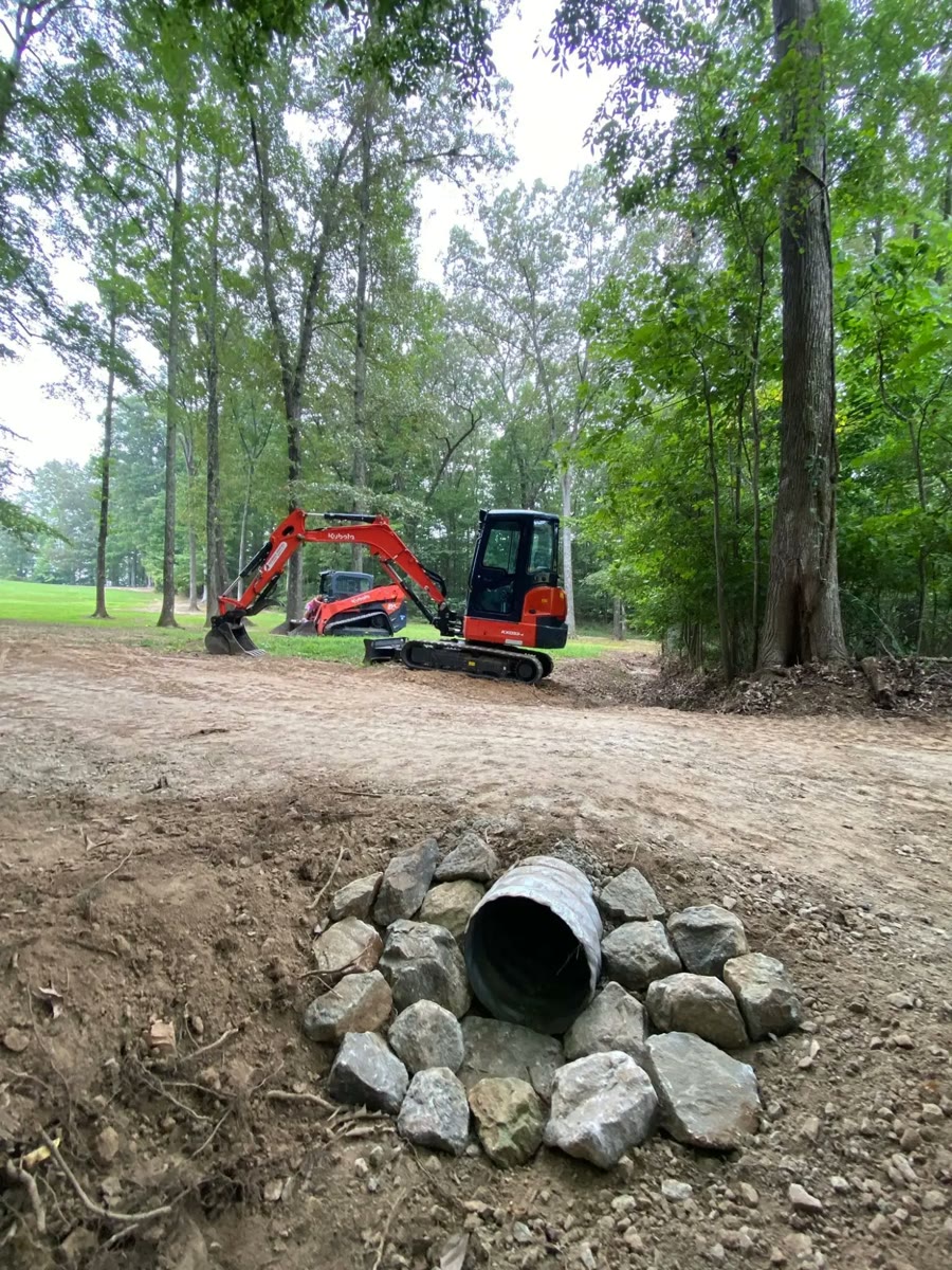 A new culvert pipe set in rock beside a dirt drive, a Kubota excavator and track loader behind it.